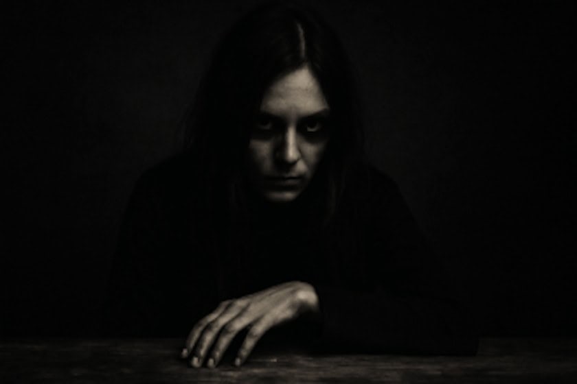 A dark, unsettling black-and-white portrait of a pale woman with shadowed eyes, staring intensely while resting her hand on a wooden surface.