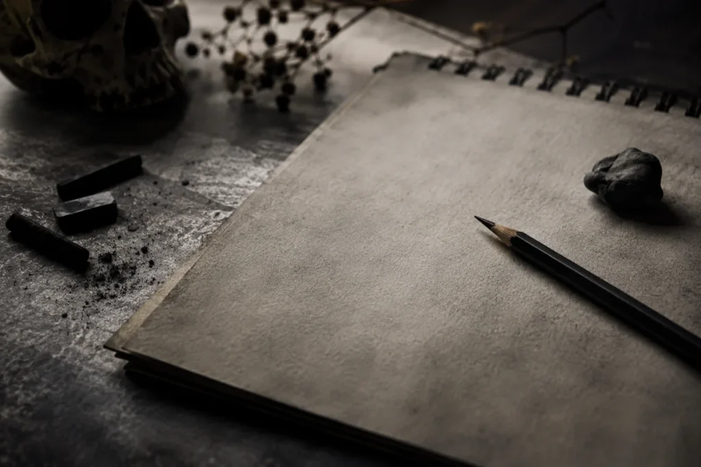 Dark atmospheric artist workspace with a blank sketchbook, graphite pencil, charcoal pieces, and subtle gothic horror elements.